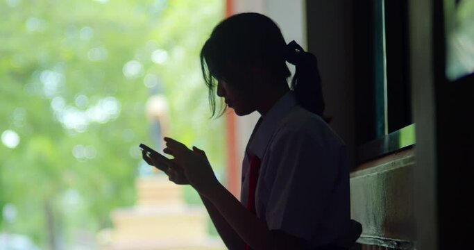A silhouette scene of a young Asian high school girl with a highly private world or introvert, casually scrolling through social media on her smartphone in the evening after school.