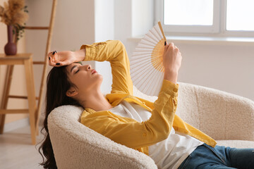 Young woman with hand fan suffering from heat on white sofa in living room