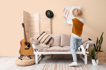 Young woman in hat hanging picture on wall in living room