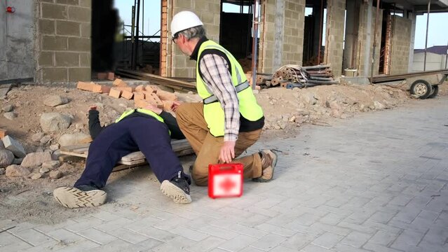 Male employee with first aid kit runs up to a fallen scaffolding worker, an injured worker, an assistant calls an ambulance, waves hand, at construction site.