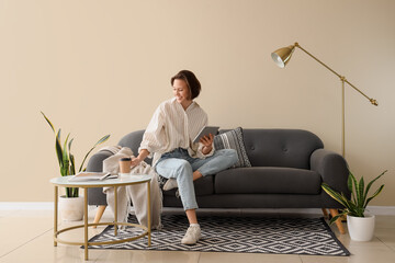 Young pretty woman using tablet and drinking coffee on sofa in light living room