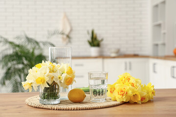 Beautiful bouquets of daffodils, glass of water and lemon on table in kitchen, closeup