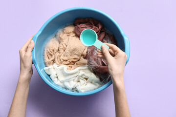Woman adding laundry detergent to clothes in plastic basin on lilac background