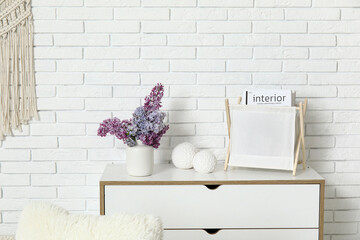 Interior of living room with bouquet of lilacs branches on chest of drawers near white brick wall