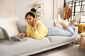 Beautiful young happy African-American woman in headphones with laptop listening music on sofa at...