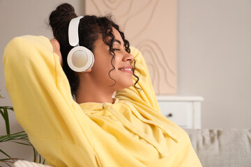 Beautiful young happy African-American woman in headphones listening music on sofa at home