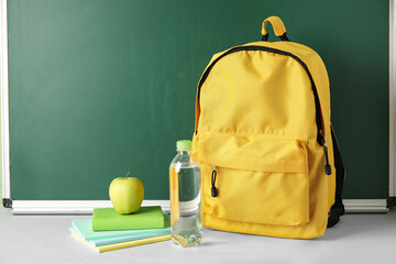 School backpack, notebooks, apple and bottle of water on table against chalkboard