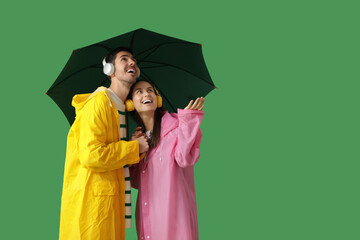Young couple in raincoats with headphones and umbrella on green background