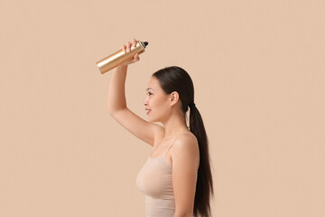 Beautiful young happy woman applying hair spray on beige background