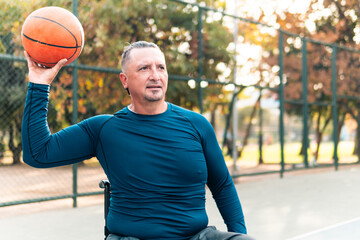 Adult male athlete about 50 years old, practicing basketball in a wheelchair, with a disability, sportswear, holding and showing the ball with joy, on a court on a sunny day