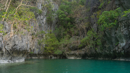 The emerald Lagoon is surrounded by sheer karst cliffs. Green tropical vegetation grows on the gray, rough slopes. Philippines. Palawan. El Nido. Small lagoon. Bacuit Bay.