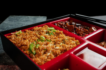 Bento box on a grunge black background with chopsticks, Containing fried rice, chicken, beef, sushi, dumplings, Chili sauce, fine dining, food photography, social media post