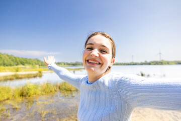 A smiling young woman stands with arms outstretched, enjoying the peaceful outdoors by a serene lake on a sunny day