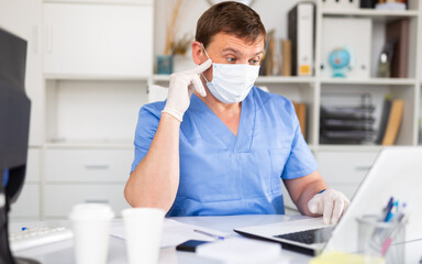 Portrait of male doctor in face mask and gloves working on laptop consulting patient online, telemedicine concept