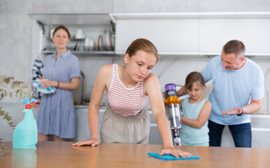 Family day at home, parents with children clean kitchen and older girl wipes dining table with rag. Dad and little girl vacuuming floor covering, mom washing dishes in background, unfocused