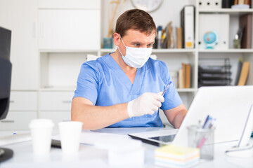 Portrait of male doctor in face mask and gloves working on laptop consulting patient online,...