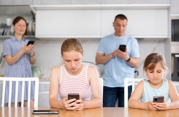 Fototapeta premium Sister girls sitting near kitchen table with phones in hands and flipping through tape and liking photos of friends, parents surf Internet in background