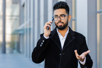 Stylish Indian businessman having remote conversation communicate speaking by smartphone outdoor. Arabian Hindu freelancer guy talking on phone standing downtown city street. Business people lifestyle