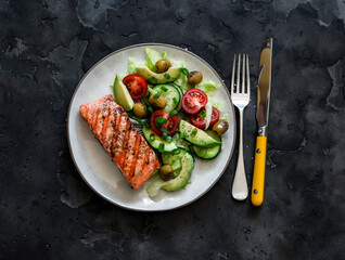 Healthy delicious lunch, dinner - grilled salmon and avocado, cherry tomatoes, cucumber, olives, iceberg lettuce on a dark background, top view
