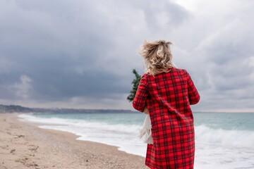 Blond woman holding Christmas tree by the sea. Christmas portrait of a happy woman walking along the beach and holding a Christmas tree in her hands. Dressed in a red coat, white dress.