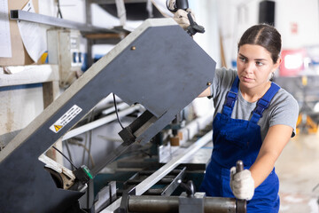 Confident young woman in blue overalls and gloves cutting PVC profile on industrial machine in...