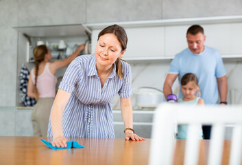 Positive woman engaged in household cleaning together with family rubbing wooden countertop in...