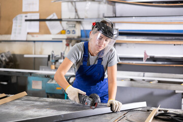 Focused skilled young woman in blue overalls, protective face shield and gloves using angle grinder...