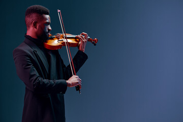 Talented young African American man in black suit playing violin with passion on dark background