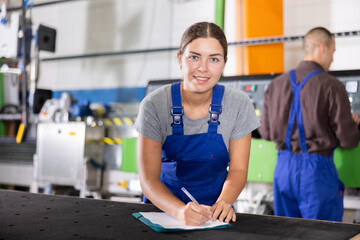 Young female glass factory worker in blue overalls smiling and taking notes on clipboard while...