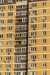A section of a multi-storey apartment block with a patterned facade with numerous windows and balconies. The exterior cladding of the building is yellow in colour with flecks of brown.