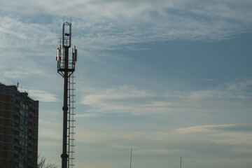 Obraz premium A telecommunication tower with broadcasting equipment, antennas and aerials rising against a clear sky. Part of a high-rise building can be seen on the right, suggesting an urban environment.