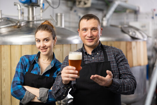 Smiling man and woman brewers presenting new beer in beer factory. Woman making thumb up gesture.