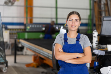 Confident smiling young female worker posing with crossed arms in glass factory, against blurred...