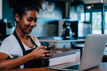 Close up portrait of focused black woman coffee shop owner in apron using smartphone in her cafe....