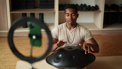 Cheerful African male blogger playing traditional ethnic music using glucophone sitting on floor in front of mobile phone with ring light. Concept of mindfulness, tranquility, harmony, sound therapy