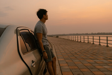 Fototapeta premium Asian man standing relaxing and leaning the car look at nature at sunset time, looking out the river, happy travel concept.