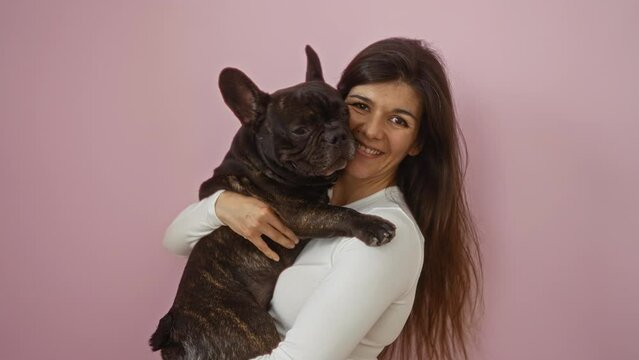 A young hispanic woman holds a french bulldog against an isolated pink background, exuding joy and warmth.