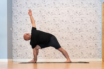 Senior man in black outfit practicing a yoga pose with one arm raised on a mat in a bright,...