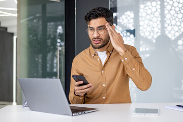 Frustrated man using phone and laptop in office, solving problem, technical issues and business stress