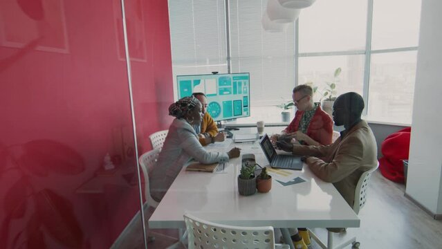 Long shot through glass wall of multiethnic project team sitting around table in meeting room, discussing business issues and planning marketing strategy for new product launch