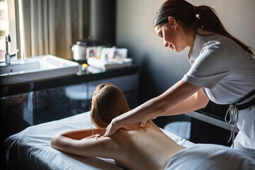 Happy professional female masseur massaging back of young relaxed woman client on massage table