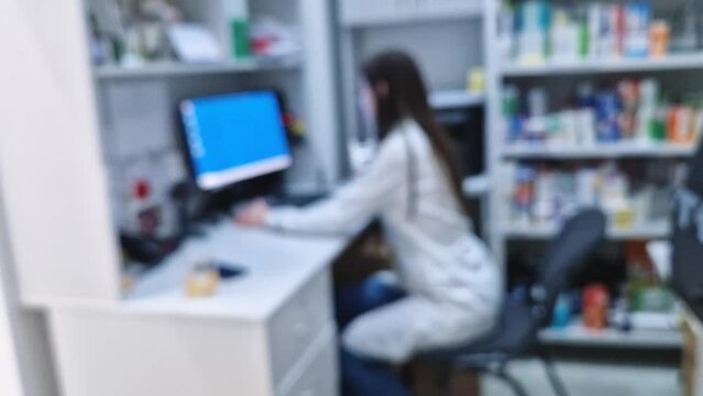Blurred view of a woman medical worker sitting in front of computer screen inside her office or laboratory. Professional female doctor in white uniform working in healthcare industry, defocus scene