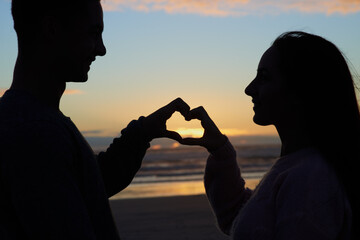 Sunset, silhouette and couple at ocean with heart hands for romance, bonding and tropical holiday...