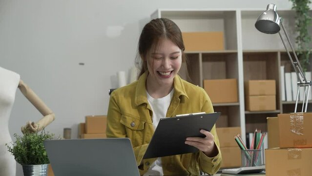 A young Asian businesswoman efficiently manages SMEs, utilizing her desk, tablet, laptop to handle online orders and live streams, showcasing entrepreneurial prowess in the dynamic market landscape.