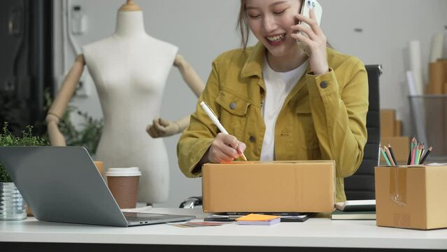 A young Asian businesswoman efficiently manages SMEs, utilizing her desk, tablet, laptop to handle online orders and live streams, showcasing entrepreneurial prowess in the dynamic market landscape.