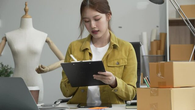 A young Asian businesswoman efficiently manages SMEs, utilizing her desk, tablet, laptop to handle online orders and live streams, showcasing entrepreneurial prowess in the dynamic market landscape.