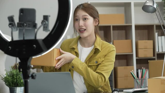A young Asian businesswoman efficiently manages SMEs, utilizing her desk, tablet, laptop to handle online orders and live streams, showcasing entrepreneurial prowess in the dynamic market landscape.