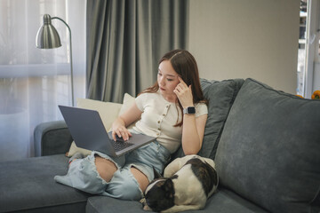 Asian teen girl uses laptop while sitting on the couch with her cute dog