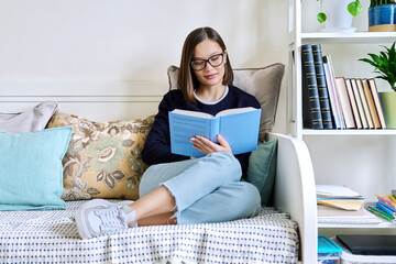 Young woman reading book, sitting on couch at home, engrossed in fiction