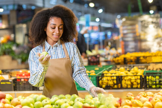 Smiling woman in apron sorting fresh apples at a grocery store, showcasing a vibrant produce section